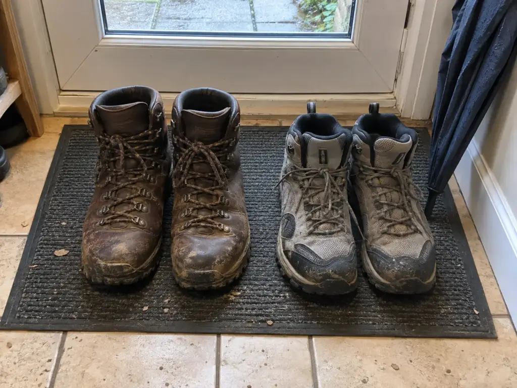 Two worn pairs of boots, one dark leather and one grey trail pair, standing on a doormat inside a back door