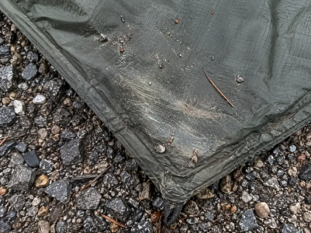 Scuffed corner of a backpacking tent floor with gravel grit stuck to the nylon after a night on rough ground