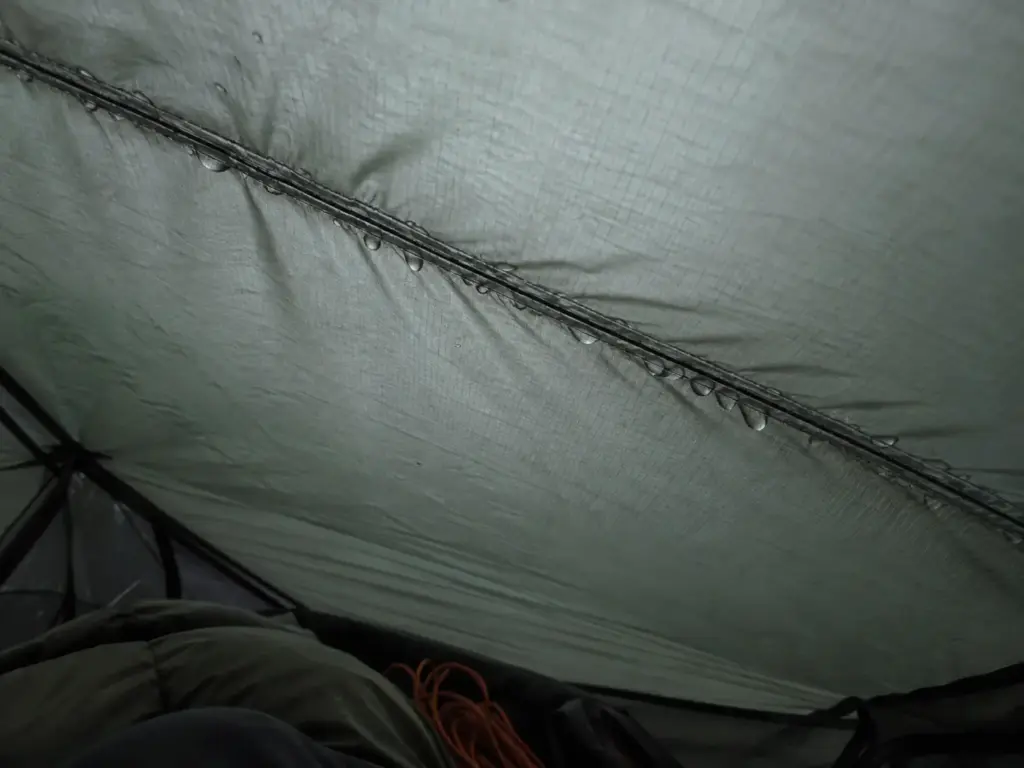 Water beads along the stitched ceiling seam of a backpacking tent after rain, a possible sign it needs seam sealing
