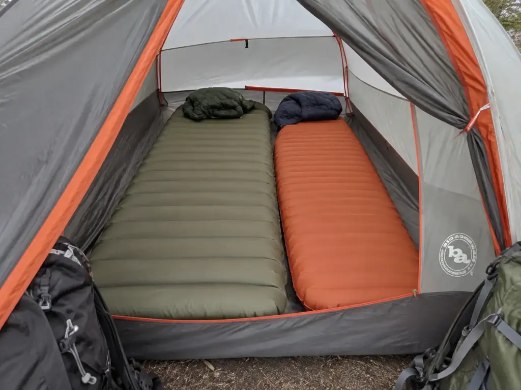 Two sleeping pads of different widths side by side on the floor of a two-person backpacking tent, seen from the open door