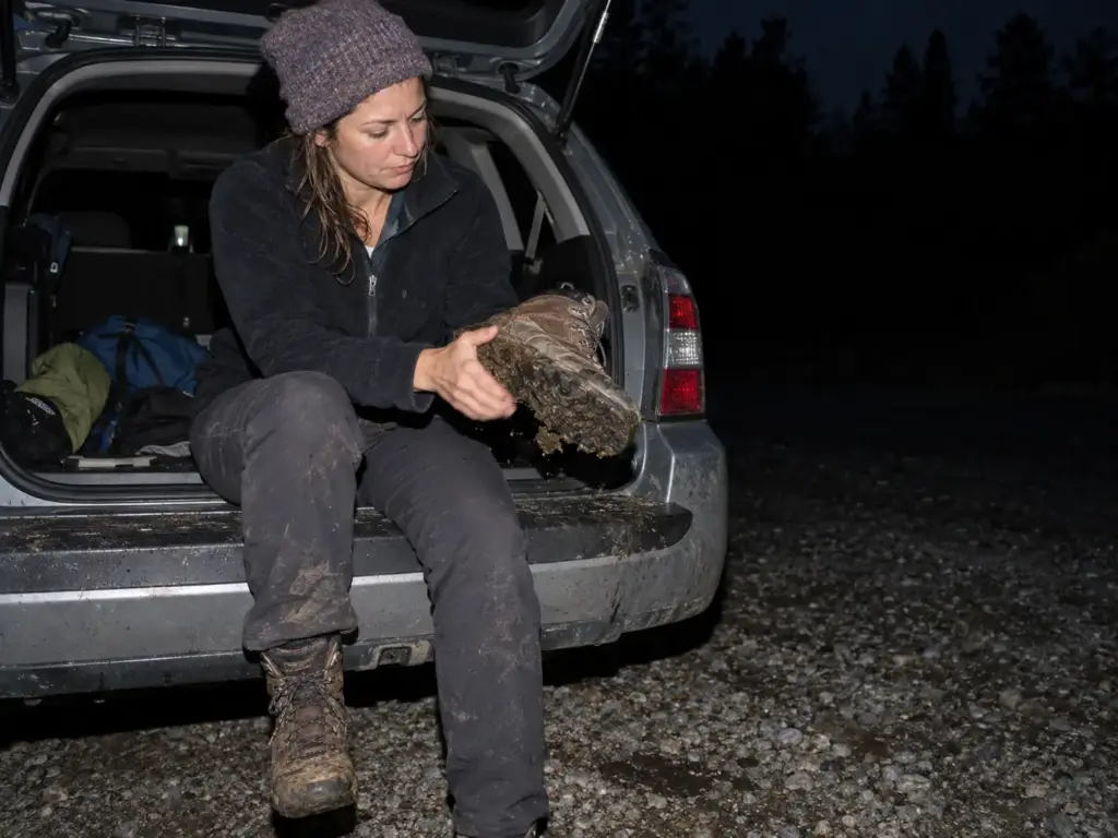Woman knocking clay off a muddy trail boot on a hatchback tailgate in a trailhead parking lot at dusk