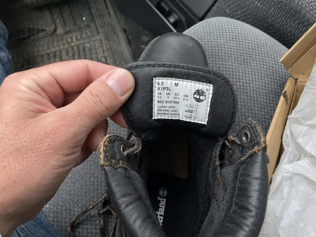 Tongue of a black steel-toe hiking boot folded forward to show the sewn-in safety label, held by a thumb