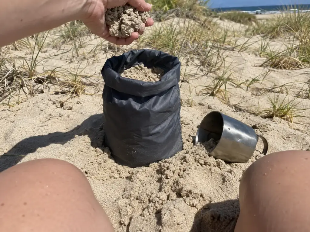 Nylon stuff sack half full of sand beside a camp cup on a beach, a quick improvised anchor for a tent in sandy soil