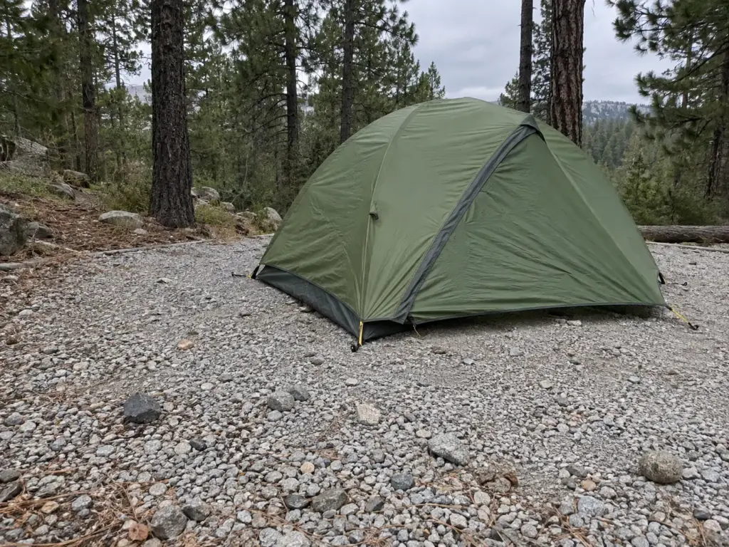 Backpacking tent pitched on a bare gravel campsite pad with half-sunk stones and a thin edge of pine needles