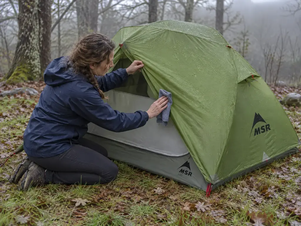 Woman wiping condensation from the inside of a double-wall backpacking tent door on a damp morning at camp