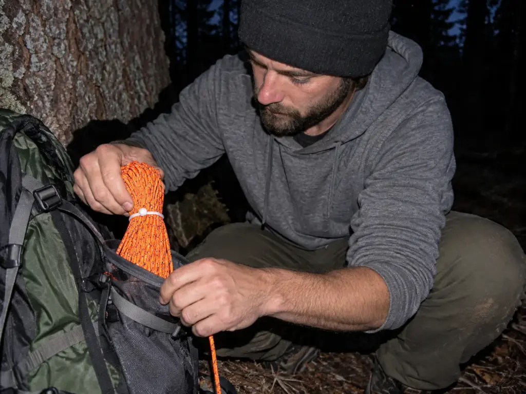 Hiker pulling a coil of reflective guyline cord from a pack at dusk, lit by on-camera flash beside a conifer