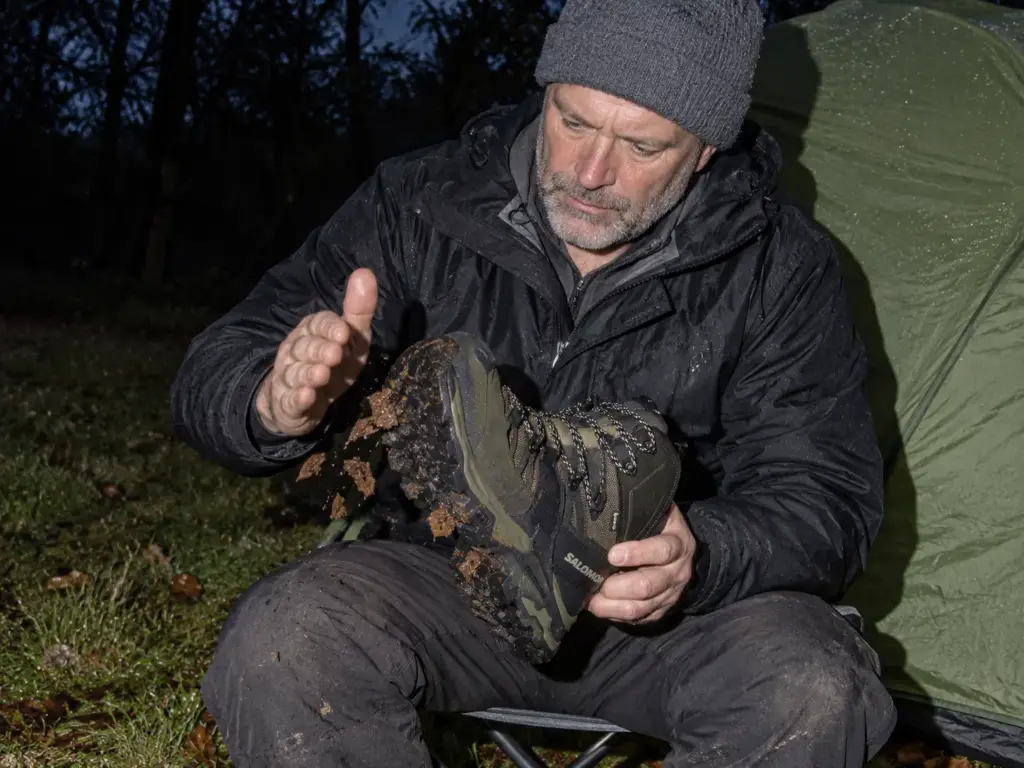Man in his 50s checking his Salomon Quest 4 GTX waterproof hiking boots beside a tent at a rainy backcountry campsite
