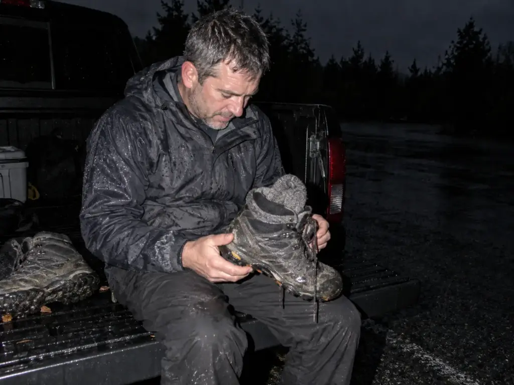 Man on a pickup tailgate at dusk tilting a soaked lightweight hiking boot to check how much water it holds