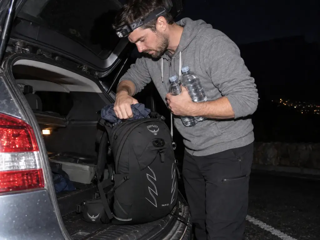 Man packs water bottles and a rain layer into an Osprey Talon 22 daypack at a car boot before sunrise on Table Mountain