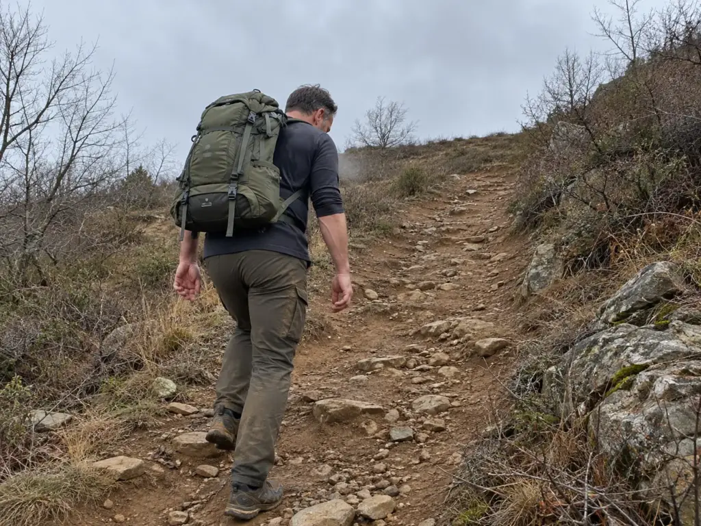 Hiker with a loaded daypack climbing a steep dirt hill trail on a grey morning, training before a Patagonia trip