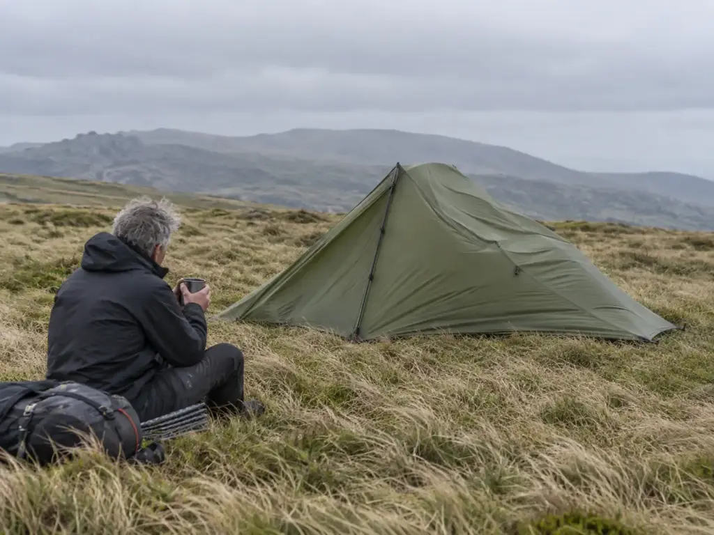 Trekking-pole tent pitched on an open meadow with its fly rippling in the wind while a camper sits nearby