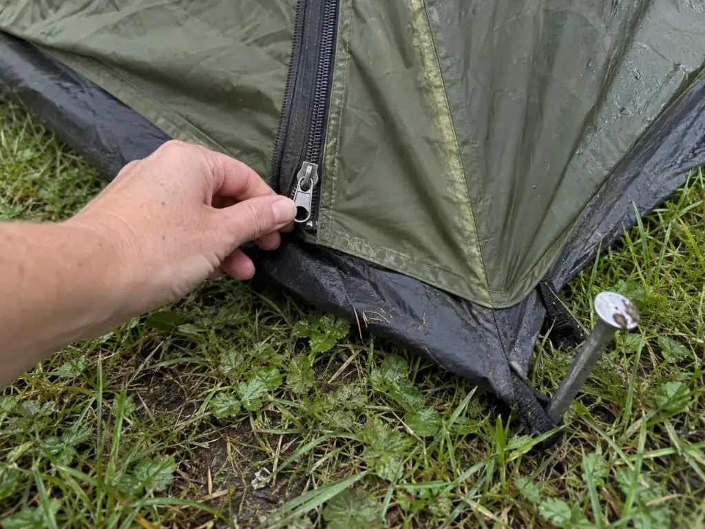 Close photo of a tent door zipper pull, polyester fly fabric and floor edge beside a stake at a campsite