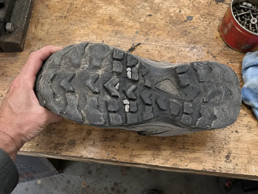 Hand steadying a lightweight hiking boot sole-up on a garage workbench to show rounded wear on the outsole lugs