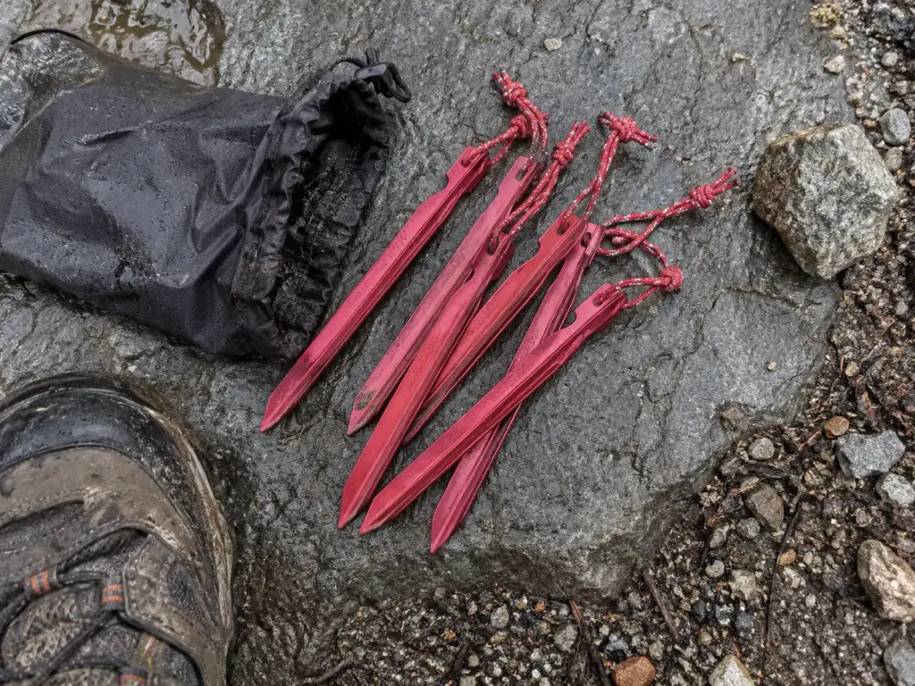 Y-beam tent stakes and their pouch on a wet grey rock beside a boot toe at a trail campsite