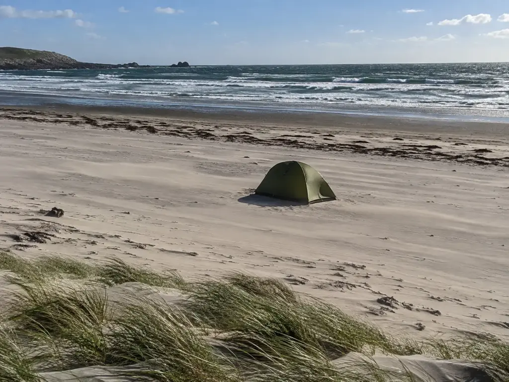 Lone dome tent on a beach flat with a darker high tide line behind it and wind-blown sand streaming across the surface