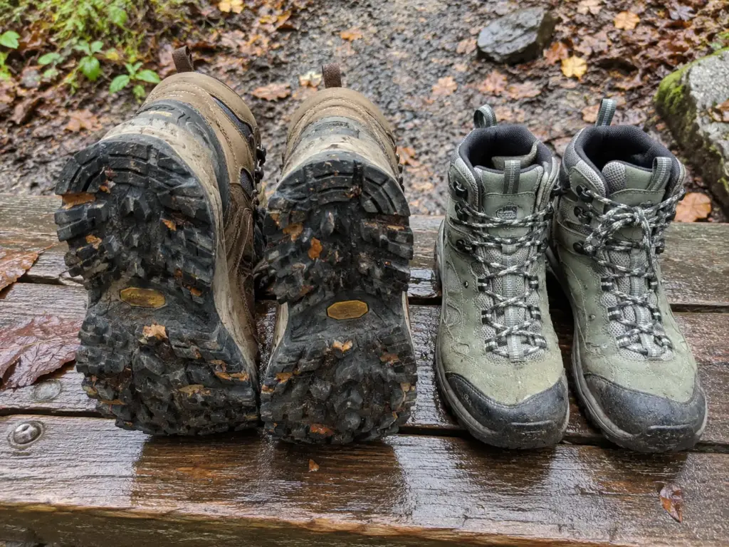 Men's and women's waterproof hiking boots side by side on a wet trailhead bench showing the lugged outsoles