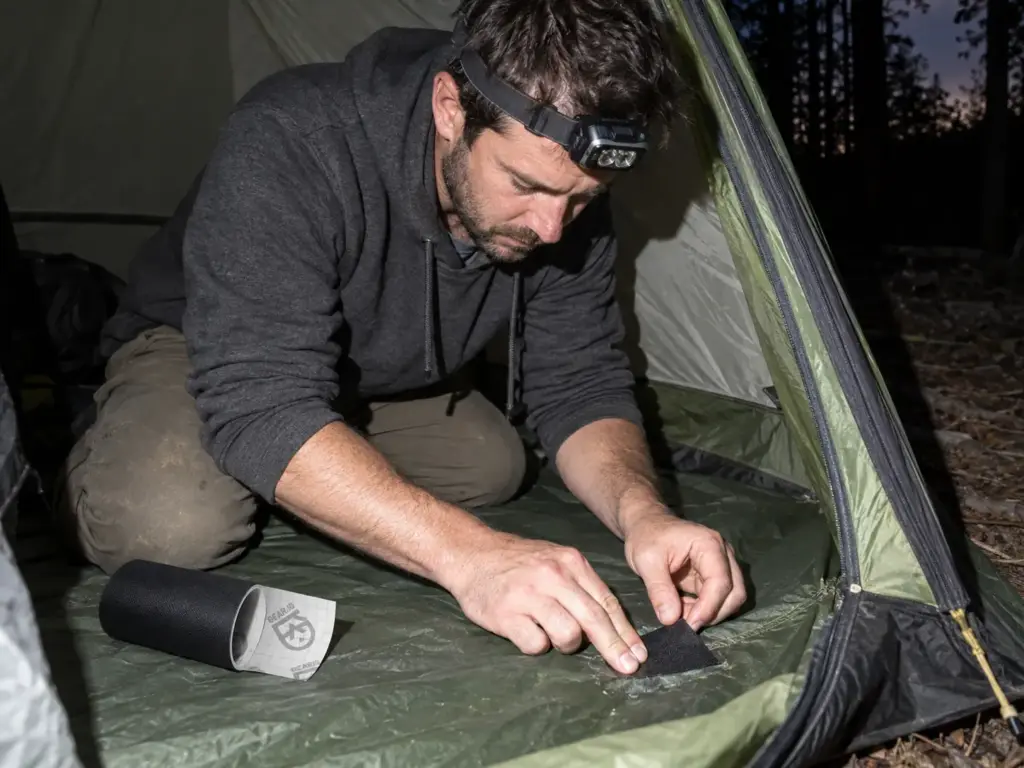 Man pressing a patch of Gear Aid Tenacious Tape onto a worn corner of a tent floor in the vestibule at dusk