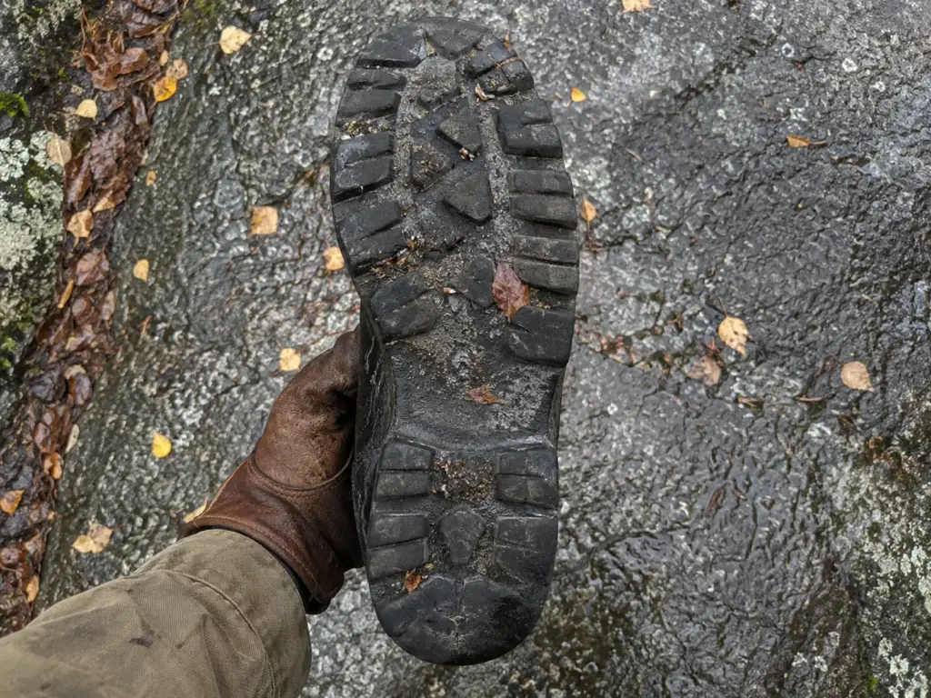 Gloved hand holding a black steel-toe work boot sole-up over a wet grey granite slab beside a trail, lugs facing the camera