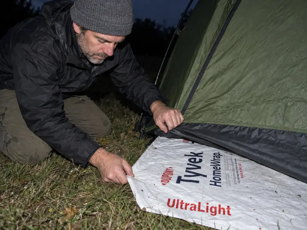 Man at dusk tucking a white Tyvek ground sheet under the floor edge of a dome tent, a cheaper footprint alternative