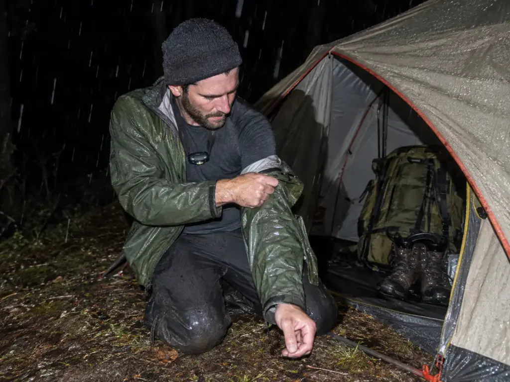 Man in his 30s at the vestibule of a pitched tent at night peeling off a wet rain shell, pack and boots inside