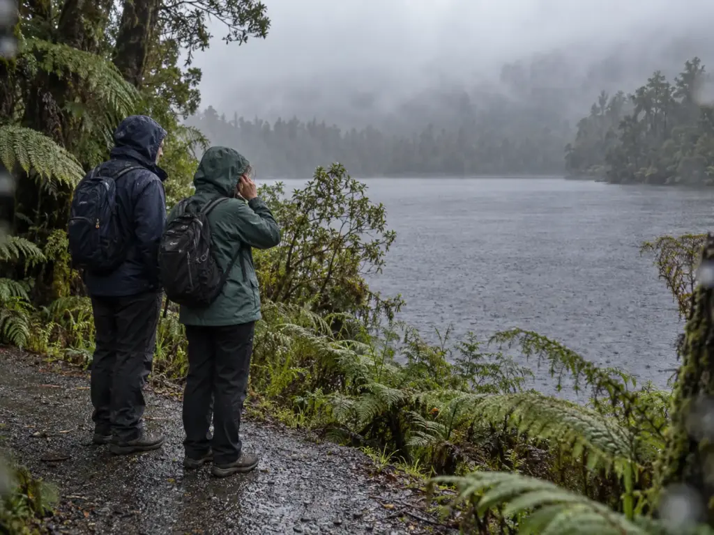 Couple in rain jackets pausing on the path beside tea-coloured Lake Matheson in West Coast rainforest, New Zealand