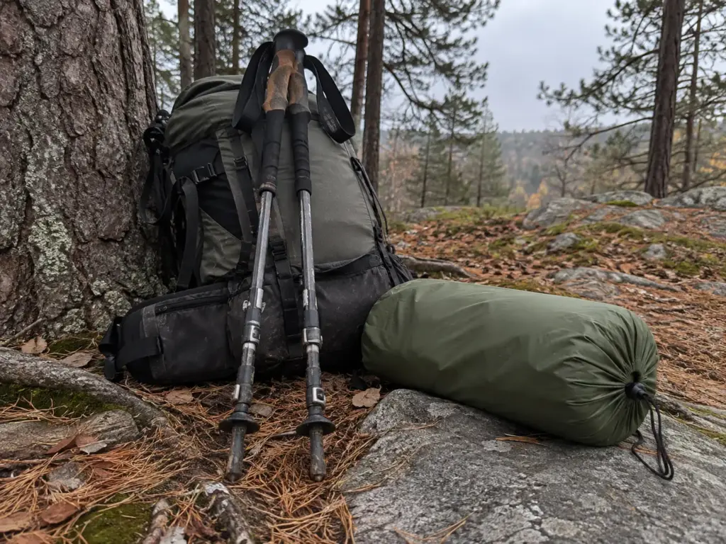 Collapsed trekking poles leaning on a loaded pack beside a tent stuff sack on a flat rock at the foot of a pine